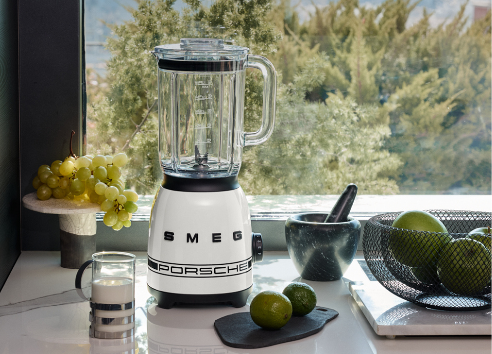Blender SMEG X Porsche collection Blanc Carrara au style années 50, présenté sur une table devant une fenêtre et entouré de récipients remplis de fruits.