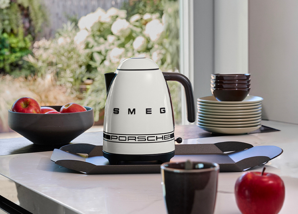Bouilloire électrique SMEG X Porsche collection Blanc Carrara 1,7L au style années 50 avec poignée chromée, présentée sur une table entourée d’assiettes, de bols et d’un saladier rempli de pommes.