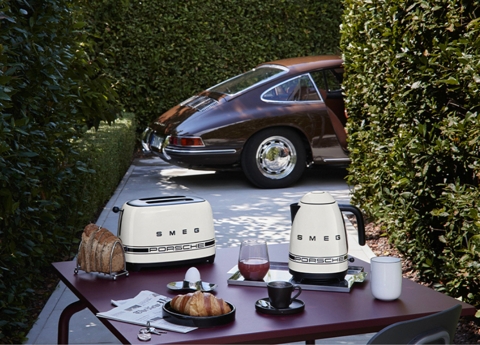 Ensemble de petit électroménager Porsche x SMEG de la collection Carrara White, composé d’une bouilloire et d’un grille-pain, posés sur une table en extérieur pour un petit-déjeuner avec croissant, jus de fruits et tasse. La table et la voiture bordeaux en arrière-plan sont entourées de buissons.