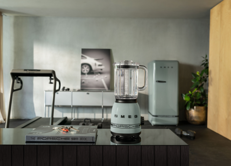 Blender Smeg X Porsche collection Blanc Carrara au style années 50 avec poignée et bec verseur noirs, présenté sur un meuble avec un livre Porsche à côté. En fond, on y voit un tapis de course, un tableau avec une voiture et sur la droite le réfrigérateur de la collection Porsche x Smeg vert ombre.