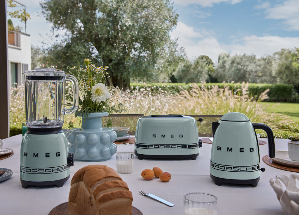 Ensemble de petit électroménager Porsche x SMEG de la collection Vert Ombre, composé d’une bouilloire, d’un blender et d’un grille-pain, disposés sur une table recouverte d’une nappe blanche dans un jardin pour un petit-déjeuner.