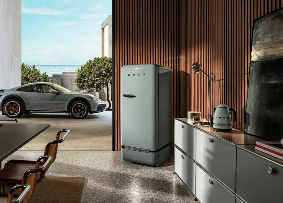 Réfrigérateur Smeg X Porsche collection Vert Ombre au style années 50 avec poignée noire, présenté dans une salle à manger ouverte sur une cour avec une voiture Porsche. Dans la salle à droite, on retrouve la bouilloire de la même collection posée sur un meuble.
