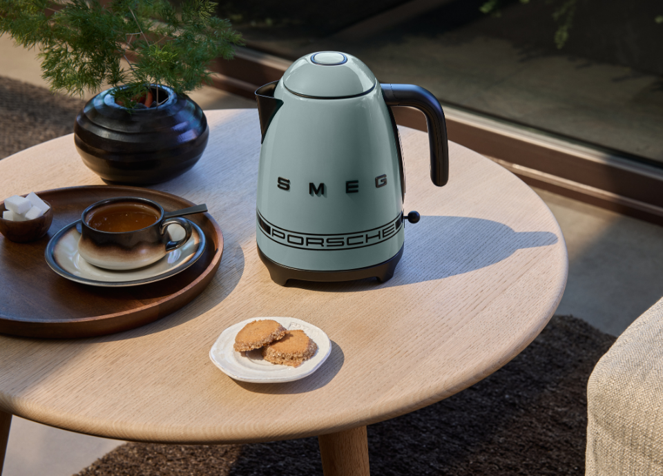 Bouilloire électrique SMEG X Porsche collection Vert Ombre 1,7L au style années 50 avec poignée et bec verseur noirs, présentée sur une table basse entourée d’une plante, d'un plateau et d'une petite assiette avec des biscuits.