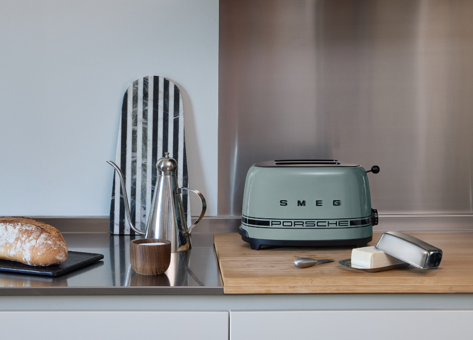 Grille-pain 2 tranches SMEG X Porsche collection Vert Ombre au style années 50 avec poignée et bec verseur noirs, présenté sur un meuble de cuisine entouré d'une théière, d'une plaquette de beurre et d'un pain.