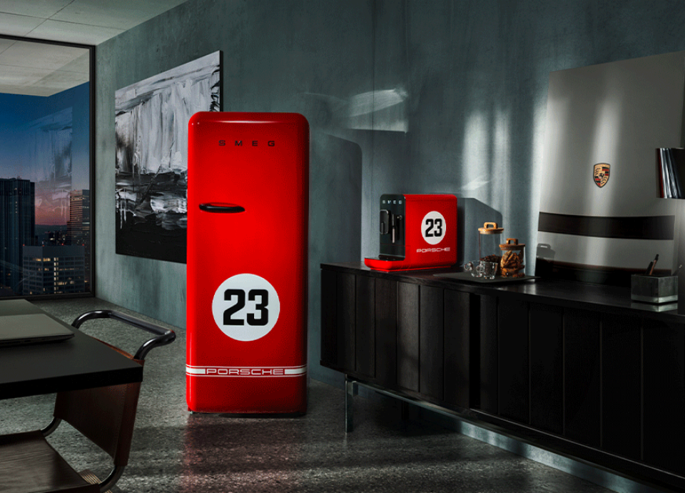 Ensemble de petit électroménager Porsche X Smeg Edition limitée 917 Salzburg au style années 50, de couleur rouge et noire, composé d’une machine à café et d’un réfrigérateur, disposés dans un salon avec vue sur une ville à travers une fenêtre située à gauche. La machine à café est posée sur un meuble haut à droite, entourée d’un plateau et d’un tableau avec le logo Porsche. À gauche, on retrouve le coin d’une table à manger avec une chaise noire.