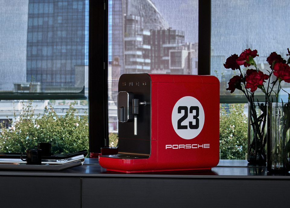 Machine à café automatique Smeg X Porsche Edition limitée 917 Salzburg au style années 50, de couleur rouge et noire avec des contours chromés sur le devant, présentée sur un meuble avec vue sur des immeubles. À droite de la machine, on retrouve un bouquet de roses tandis qu’à sa gauche se trouve un plateau avec des tasses.