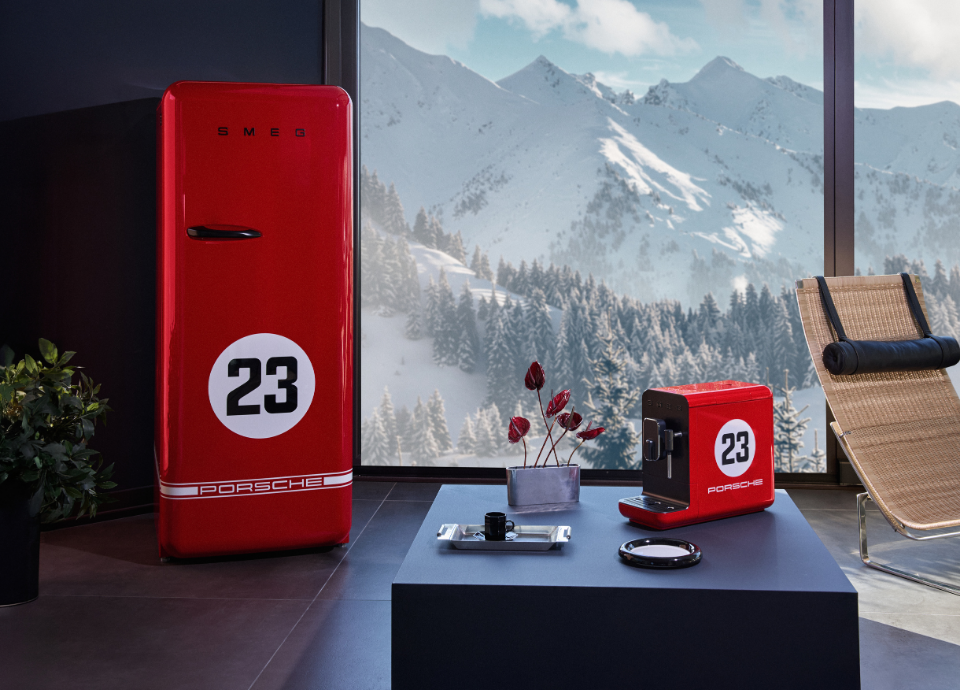 Ensemble de petit électroménager Porsche X Smeg Edition limitée 917 Salzburg au style années 50, de couleur rouge et noire, composé d’une machine à café et d’un réfrigérateur, disposés dans un salon avec vue sur des montagnes enneigées. Le réfrigérateur se trouve à gauche, tandis que la machine à café est posée sur une table basse noire, entourée d’une plante et d’un plateau. À droite de l’image, on retrouve une chaise en osier.