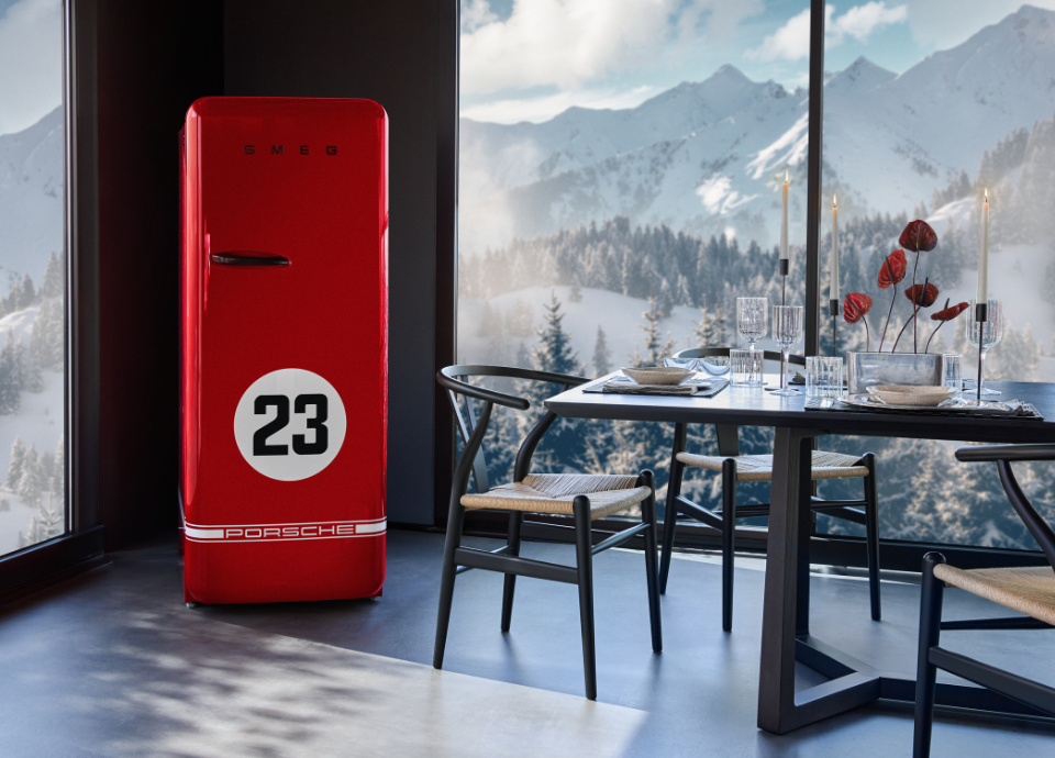 Réfrigérateur Smeg X Porsche Edition limitée 917 Salzburg au style années 50 avec poignée noire et revêtement rouge, présenté dans une salle à manger ouverte avec vue sur des montagnes enneigées. Dans la salle, on retrouve une table à manger avec des chaises noires et une table dressée avec des chandeliers.