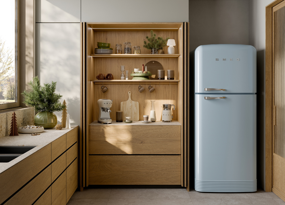 Réfrigérateur deux portes Smeg style années 50 en coloris bleu pastel avec poignées chromées, doté d’un compartiment congélateur en partie supérieure et d’un réfrigérateur en partie inférieure. Il est installé dans une cuisine contemporaine aux finitions en bois naturel.  À ses côtés, une niche de rangement ouverte avec étagères en chêne présente de la vaisselle, des bocaux, des accessoires de décoration et plusieurs petits appareils électroménagers Smeg. Au premier plan à gauche, un plan de travail en pierre claire agrémenté d’un vase végétal et d’éléments décoratifs complète l’ambiance chaleureuse et lumineuse de la pièce.