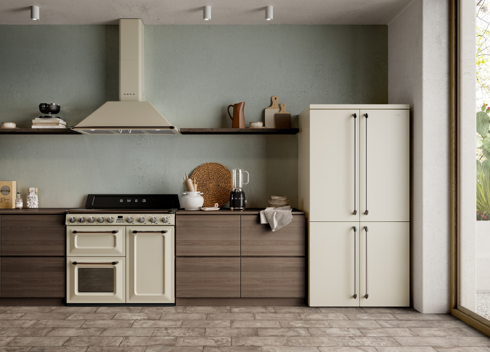 Blender Smeg Années 50 en finition noir brillant posé sur le plan de travail, accompagné d’un piano de cuisson Smeg esthétique Victoria crème avec crédence noire et manettes métalliques. Au-dessus, une hotte murale assortie de la collection Victoria complète l’ensemble. À droite, un réfrigérateur Smeg crème au style rétro s’intègre dans une cuisine aux façades effet bois foncé, avec étagères décoratives et mur vert sauge.