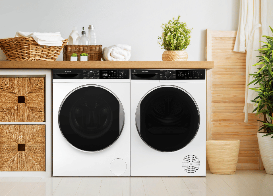 Lave-linge et sèche-linge SMEG installés côte à côte dans une buanderie lumineuse au style naturel. Entourés de paniers en osier, de linge soigneusement plié et de plantes décoratives, les appareils illustrent un soin du linge optimal pour des vêtements prêts à porter dès la sortie du sèche-linge.