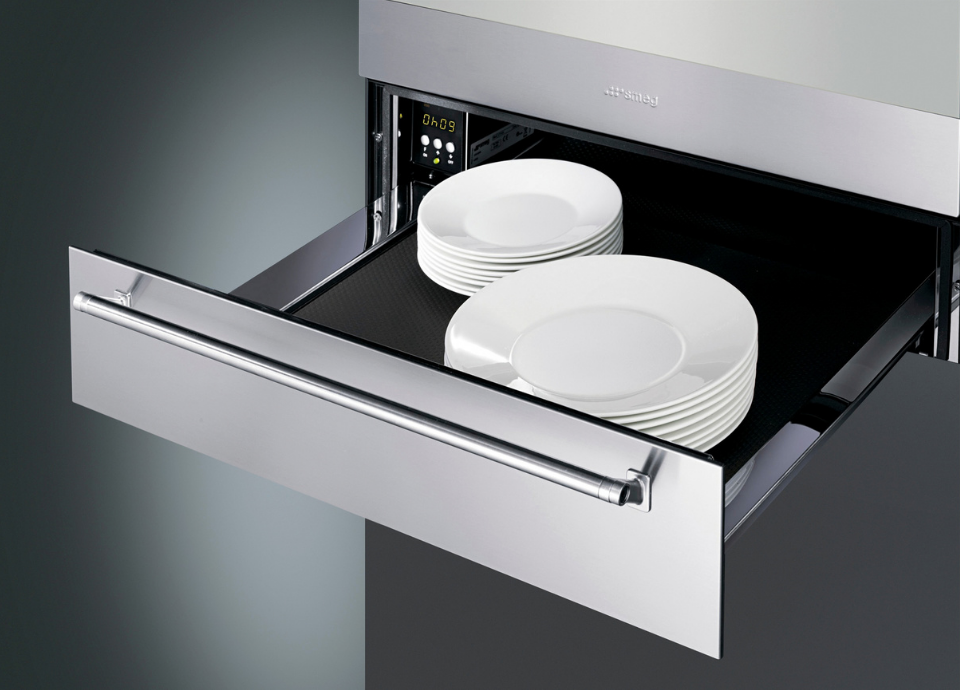 Tiroir chauffant Smeg en inox avec intérieur noir, présenté ouvert pour dévoiler plusieurs assiettes rangées à l’intérieur. Des boutons de réglage sont situés au fond à gauche du tiroir pour le contrôle des fonctions. Sur la partie supérieure de la façade se trouve le logo Smeg, tandis qu’une poignée arrondie s’étend sur toute la largeur du tiroir. Son design élégant s’intègre parfaitement dans une cuisine moderne haut de gamme.