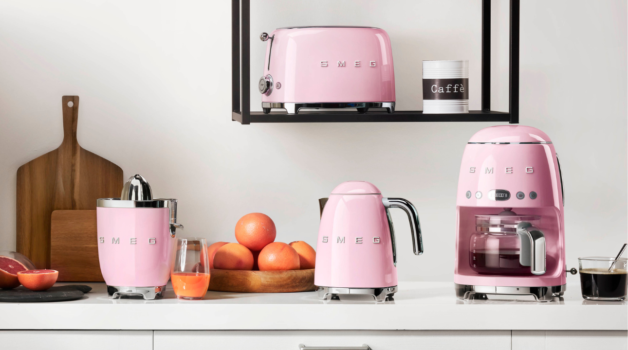 Ensemble de petit-déjeuner SMEG machine à café filtre rose, bouilloire rose, grille-pain rose et presse-agrumes rose sur un plan de travail.