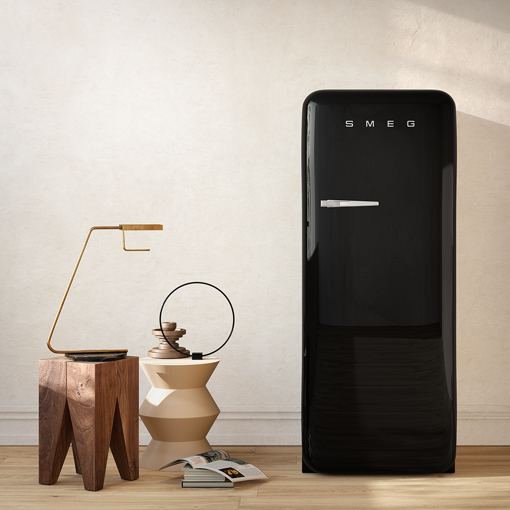 Un réfrigérateur Smeg noir aux finitions laquées et aux angles arrondis emblématiques du style vintage. Il est placé dans un intérieur moderne et épuré. À sa gauche, on trouve deux tables d'appoint sculpturales (une en bois brut, l'autre géométrique beige) surmontées d'une lampe dorée minimaliste et d'un objet décoratif circulaire noir. Quelques magazines sont posés au sol sur un parquet clair, devant un mur texturé crème.