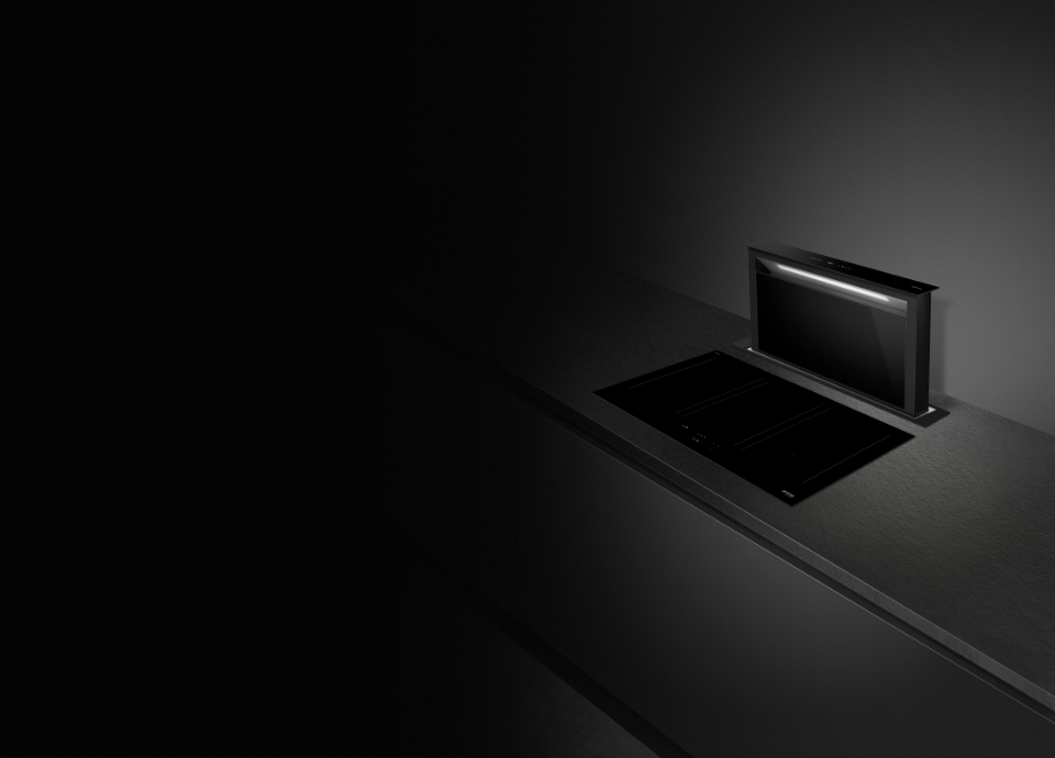 Hotte de plan Smeg noire, intégrée au plan de travail et ouverte en position haute. La hotte est positionnée à droite avec un éclairage mettant en valeur son design moderne et son intégration discrète dans la cuisine.