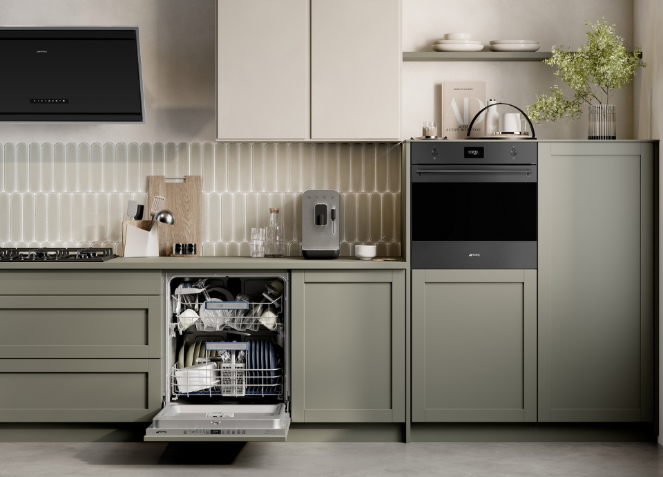Lave-vaisselle encastrable Smeg de 60 cm intégré dans une cuisine moderne de couleur kaki. Le lave-vaisselle ouvert est rempli de couverts. Sur le plan de travail se trouvent une gazinière, une machine à café, une hotte Smeg et divers ustensiles de cuisine. À droite, des placards de cuisine en hauteur intègrent un four Smeg. Au-dessus sont disposés une plante, un livre et des couverts.