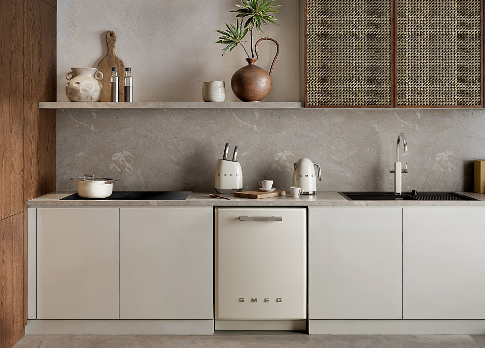 Lave-vaisselle encastrable Smeg crème au style années 50, intégré dans une cuisine de couleur crème. Sur le plan de travail se trouvent plusieurs ustensiles de cuisine Smeg, notamment une casserole, une bouilloire, un mitigeur et un bloc de couteaux. Au-dessus du plan de travail, une étagère murale accueille des vases et une planche à découper, avec un meuble en bois et osier à proximité. À gauche, un mur en bois complète la cuisine. Le plan de travail est surmonté d’une crédence en marbre gris clair.