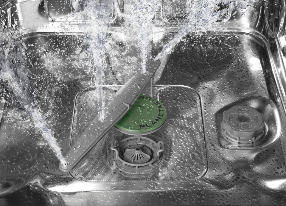 Vue intérieure d’un lave-vaisselle Smeg en inox. Le bras de lavage rotatif gris et le système Planetarium vert sont visibles au fond de la cuve. De l’eau est projetée par les jets du système Planetarium afin d’assurer une diffusion homogène dans toute la cavité du lave-vaisselle.