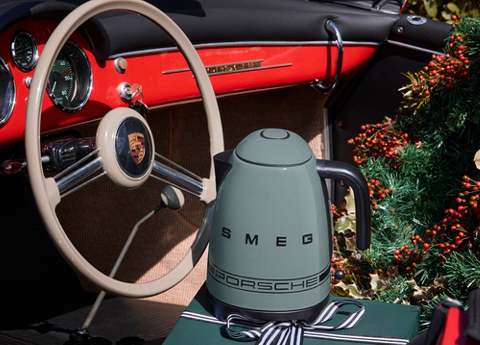 Innenraum eines klassischen Porsche-Sportwagens mit einem hellen Lenkrad und einem roten Armaturenbrett. Auf dem Sitz steht ein dunkelgrüner SMEG-Wasserkocher mit Porsche-Logo, platziert auf einer grünen Geschenkbox, die mit einem schwarz-weißen Band gebunden ist. Im Hintergrund befinden sich Weihnachtsdekorationen mit roten Beeren.