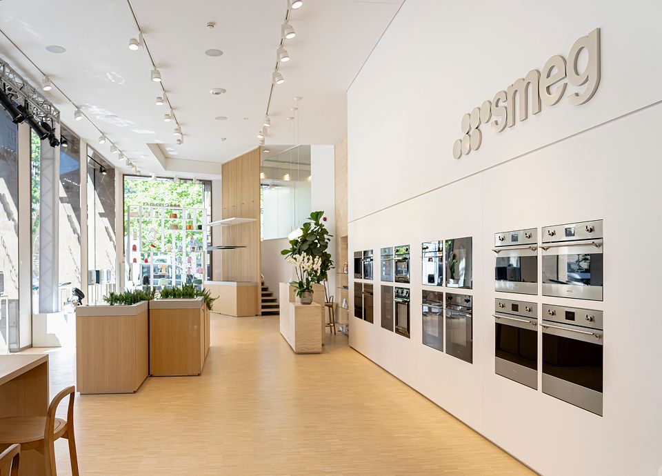 Milan SMEG store showroom with floor‑to‑ceiling windows, light wood flooring, and a wall display of built‑in stainless steel ovens beneath the Smeg logo, with plants and wooden display units arranged throughout the open space.