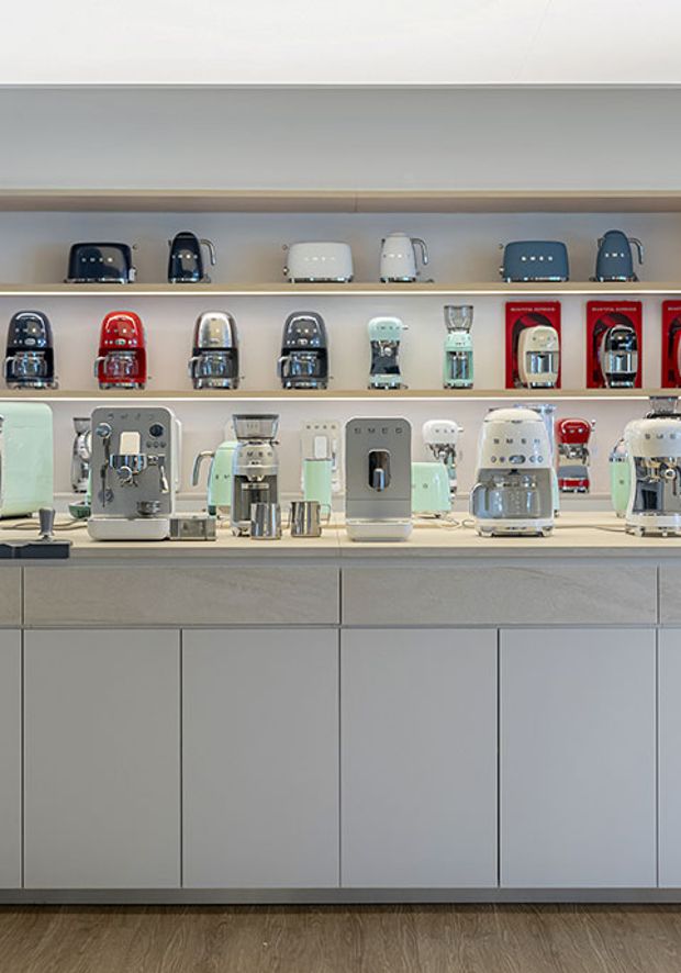 Area espositiva dei piccoli elettrodomestici presso lo SMEG Store Reggio Emilia. In primo piano sono esposte macchine per il caffè, bollitori, tostapane, frullatori e altri prodotti SMEG in diverse finiture e colori, organizzati su un'isola centrale e scaffalature dedicate per consentire ai visitatori di scoprire da vicino design e funzionalità della collezione.