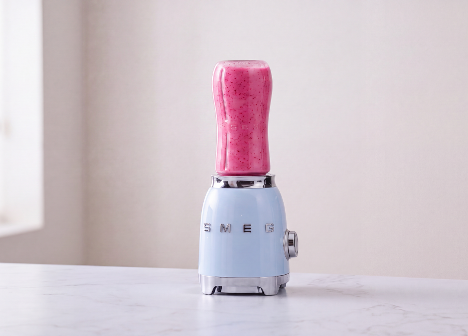 SMEG Personal Blender in crèmekleur met een roze smoothie in de drinkfles, geplaatst op een houten keukenblad. Op de voorgrond liggen twee gehalveerde kiwi's op een houten plankje, met een zachte, onscherpe keukenachtergrond.