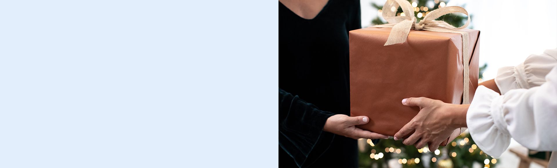 Two people exchange a large rectangular gift wrapped in brown paper and tied with a beige ribbon. A Christmas tree with warm lights is visible in the background.