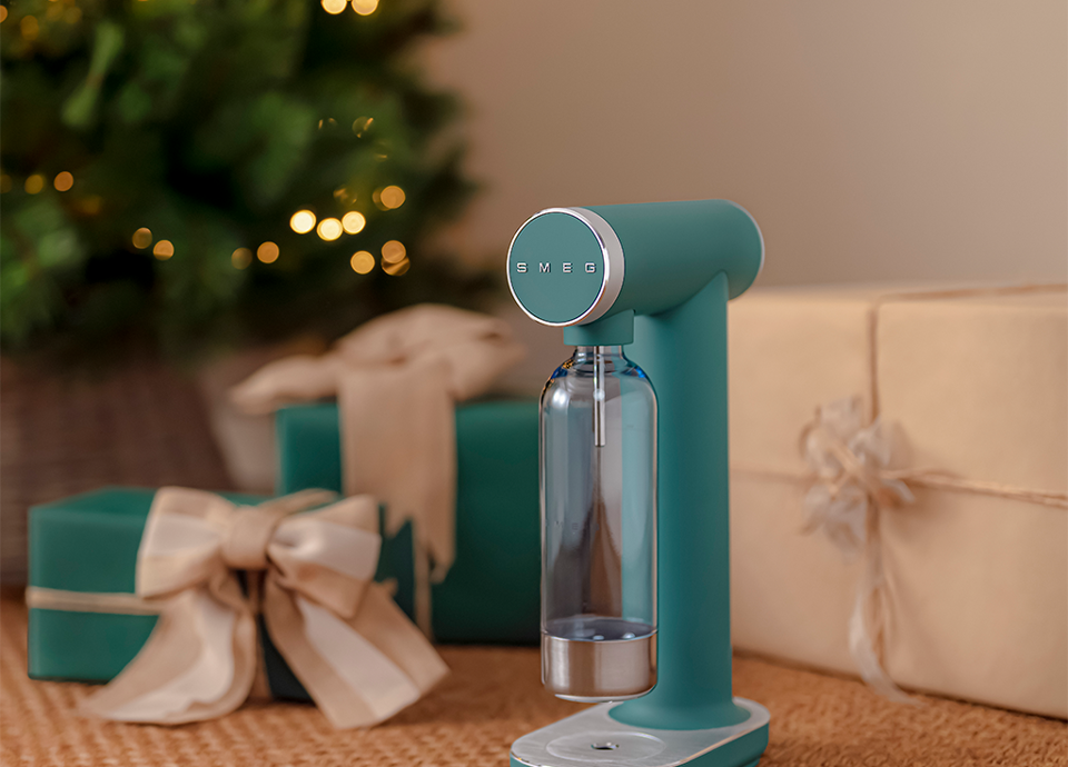 a lit Christmas tree. Emerald green SMEG water carbonator with a transparent bottle, placed on a rug in front of gift boxes wrapped in beige and green paper and decorated with bows. In the background,