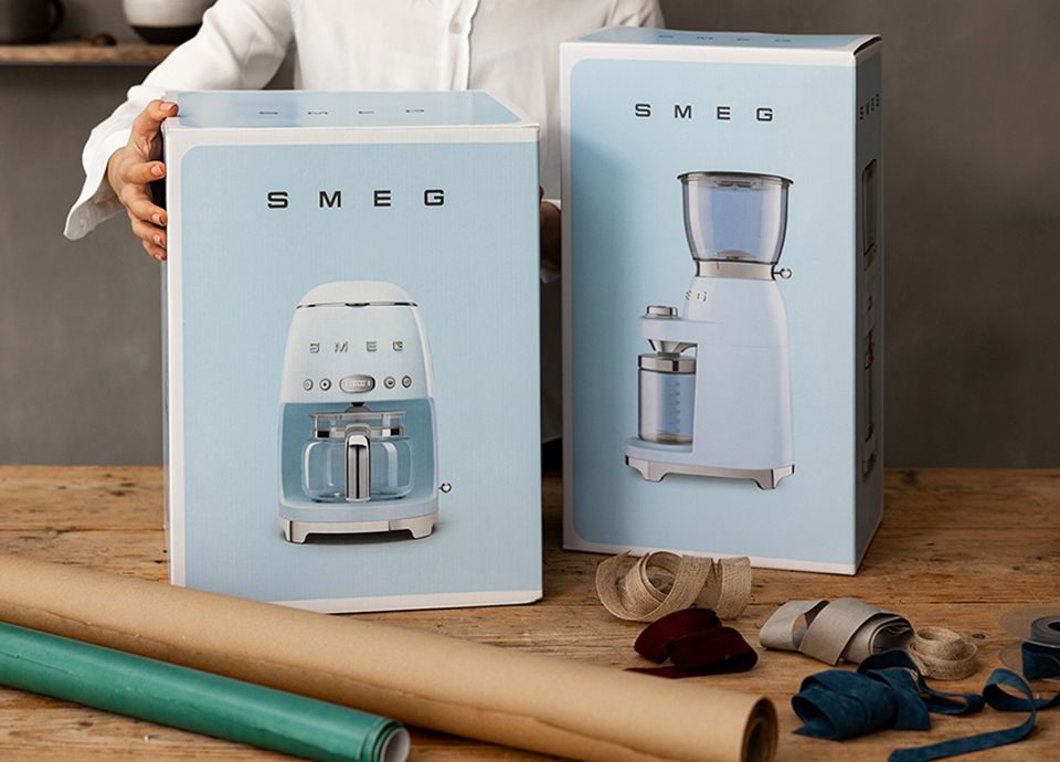 Filter coffee machine and milk frother by SMEG, both in cream color, placed on a light wooden cabinet, flanked by two gift boxes wrapped in brown paper and decorated with pine branches and red berries. In the background, a green wall with a Christmas wreath.