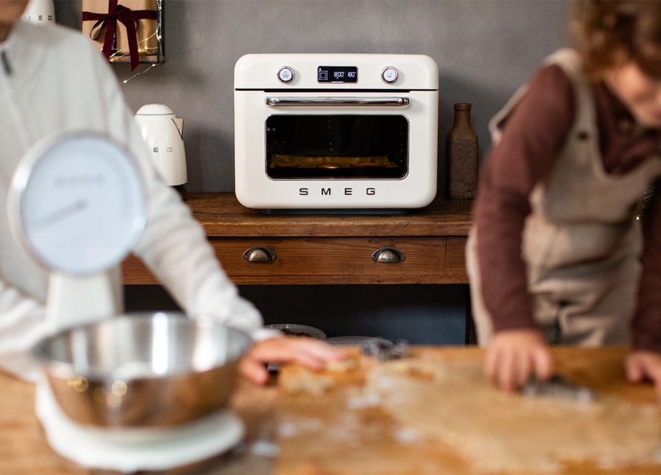 Hands holding an emerald green SMEG kitchen scale, resting on a marble countertop. Blurred decorative lights in the background.