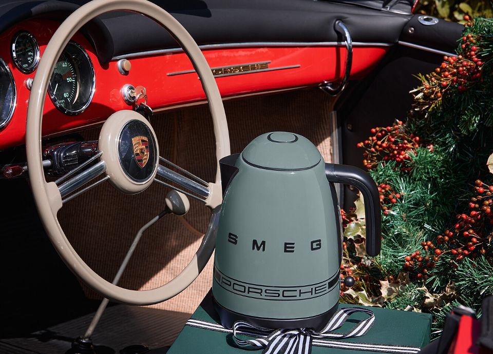 Interior of a vintage Porsche sports car with a light steering wheel and red dashboard. On the seat, there is a SMEG dark green kettle with logo Porsche, placed on a green gift box tied with a black-and-white ribbon. In the background, Christmas decorations with red berries.