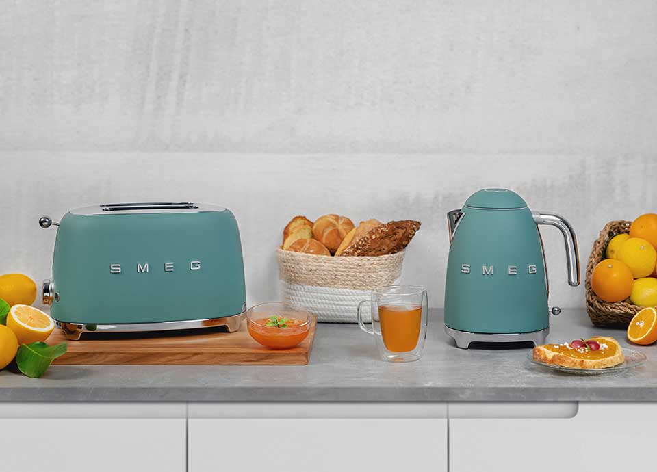 Our emerald green toaster and kettle, on a grey marble worktop, interspersed with a range of delicious looking breakfast food items including fresh bread, oranges, and a clear glass mug containing freshly brewed tea without milk.