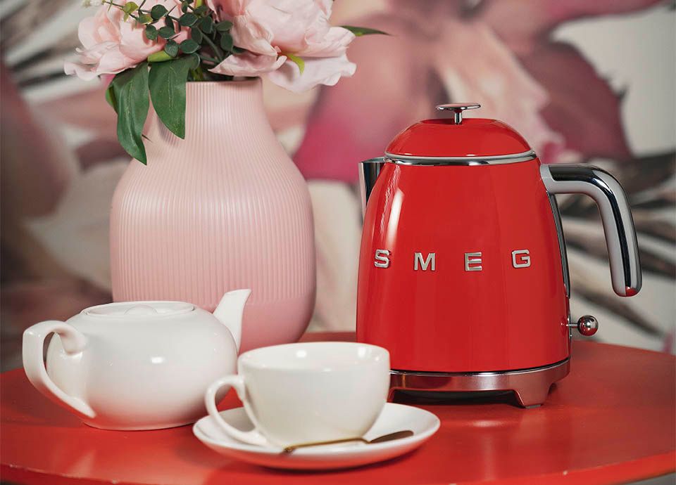 Smeg's mini kettle in red on a red table with a white tea pot, cup and saucer, and a pink vase with pink flowers