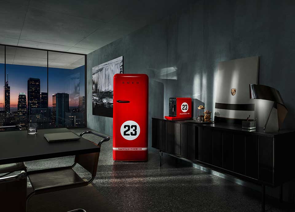 Un interno moderno ed elegante, con un'ampia finestra che rivela un paesaggio urbano al tramonto. La stanza è caratterizzata da eleganti mobili scuri e un arredamento minimalista.  In bella vista, un frigorifero SMEG rosso e una macchina da caffè rossa, entrambi contrassegnati da un cerchio bianco contenente il numero "23" e il logo "PORSCHE".  Il frigorifero presenta il marchio "SMEG" in grassetto nella parte superiore. Dietro, un dipinto astratto in bianco e nero aggiunge contrasto alla scena.  Il design enfatizza colori vivaci, linee pulite e un forte contrasto visivo, garantendo leggibilità e accessibilità per gli utenti con disabilità visive.
