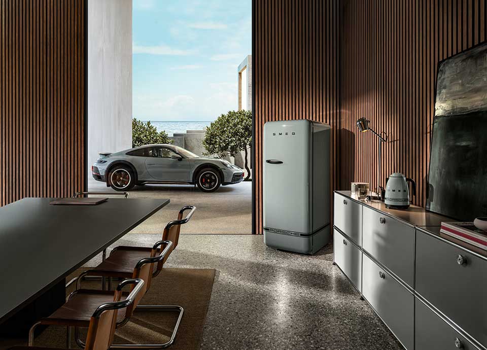 Uno spazio moderno con boiserie in legno e un lungo tavolo circondato da sedie. Su un lato della stanza, si trovano un frigorifero Porsche x SMEG Shade Green e una credenza con un bollitore Porsche x SMEG Shade Green, libri, una lampada e piccoli oggetti decorativi. Ampie finestre offrono una vista panoramica sul verde, su una Porsche 911 GT3 RS parcheggiata all'esterno e sull'oceano sullo sfondo. L'ambiente è spazioso e ben illuminato da luce naturale, garantendo una visibilità ottimale e un'accessibilità ottimale anche per gli utenti con disabilità visive.