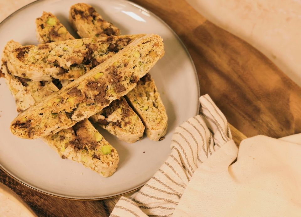 Biscotti slices with pistachio pieces arranged on a ceramic plate, styled on a wooden surface with a folded cloth.