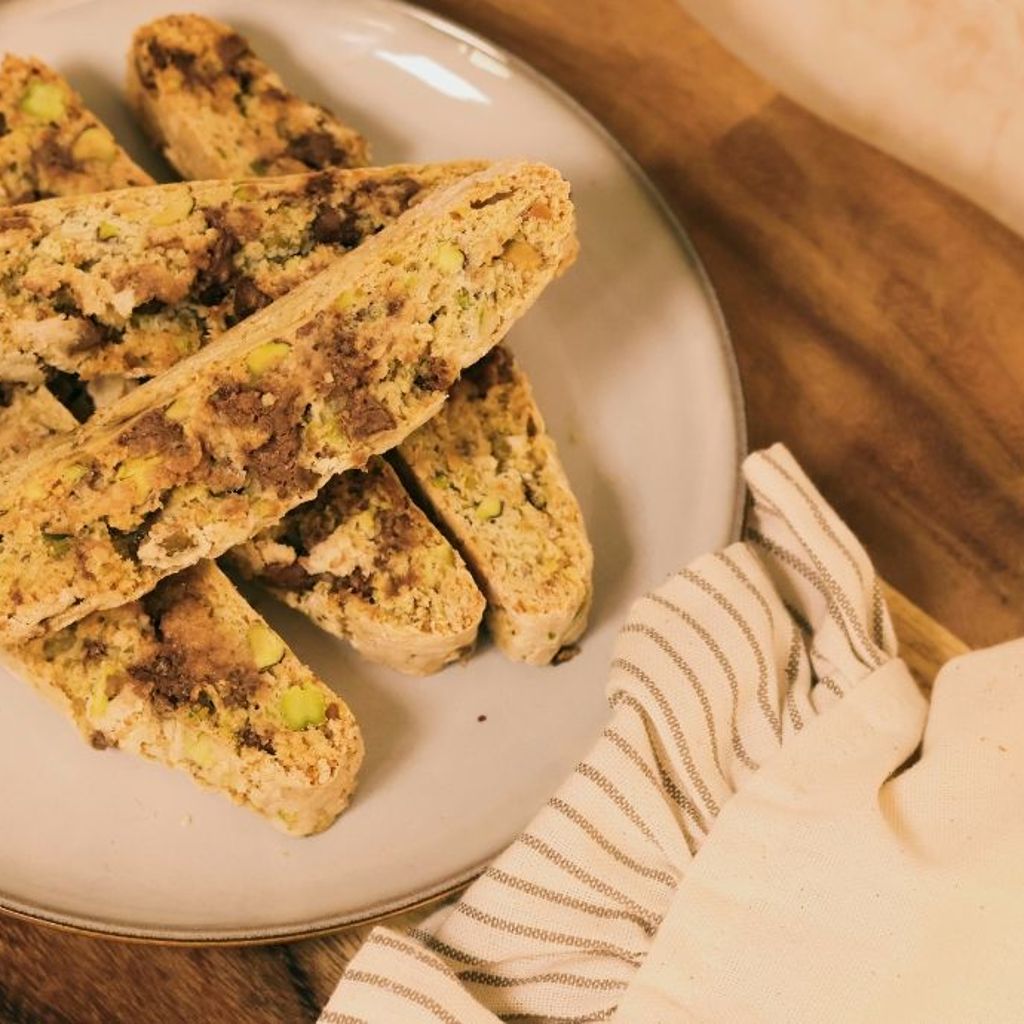 Biscotti slices with pistachio pieces arranged on a ceramic plate, styled on a wooden surface with a folded cloth.