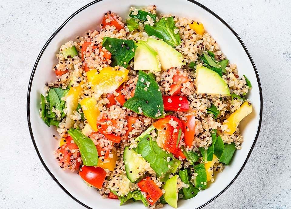 Overhead view of a vibrant quinoa salad mixed with chopped red and yellow peppers, cucumber, and spinach in a white bowl.