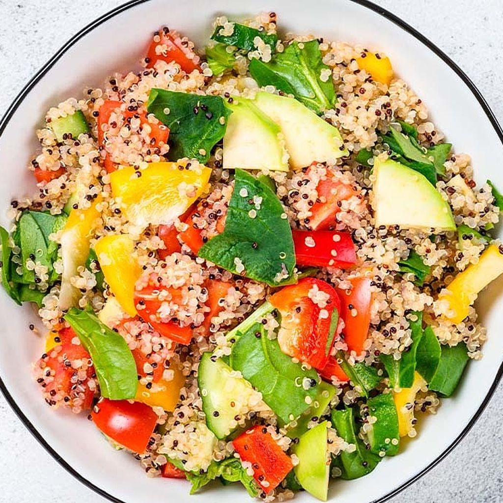 Ciotola bianca di quinoa mista a peperoni gialli e rossi, cetrioli, pomodori e spinaci freschi, vista dall'alto.