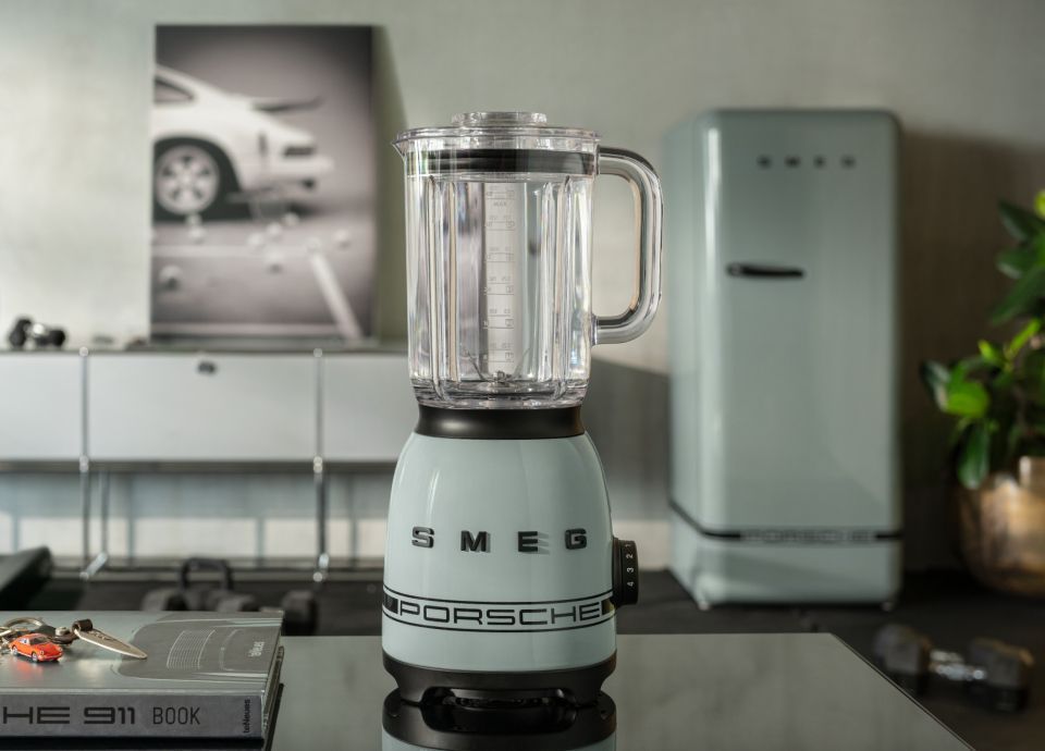 Smaragdgrüner SMEG-Wassersprudler mit einer transparenten Flasche, auf einem Teppich platziert vor Geschenkboxen in beige- und grünfarbenem Papier, verziert mit Schleifen. Im Hintergrund steht ein beleuchteter Weihnachtsbaum.