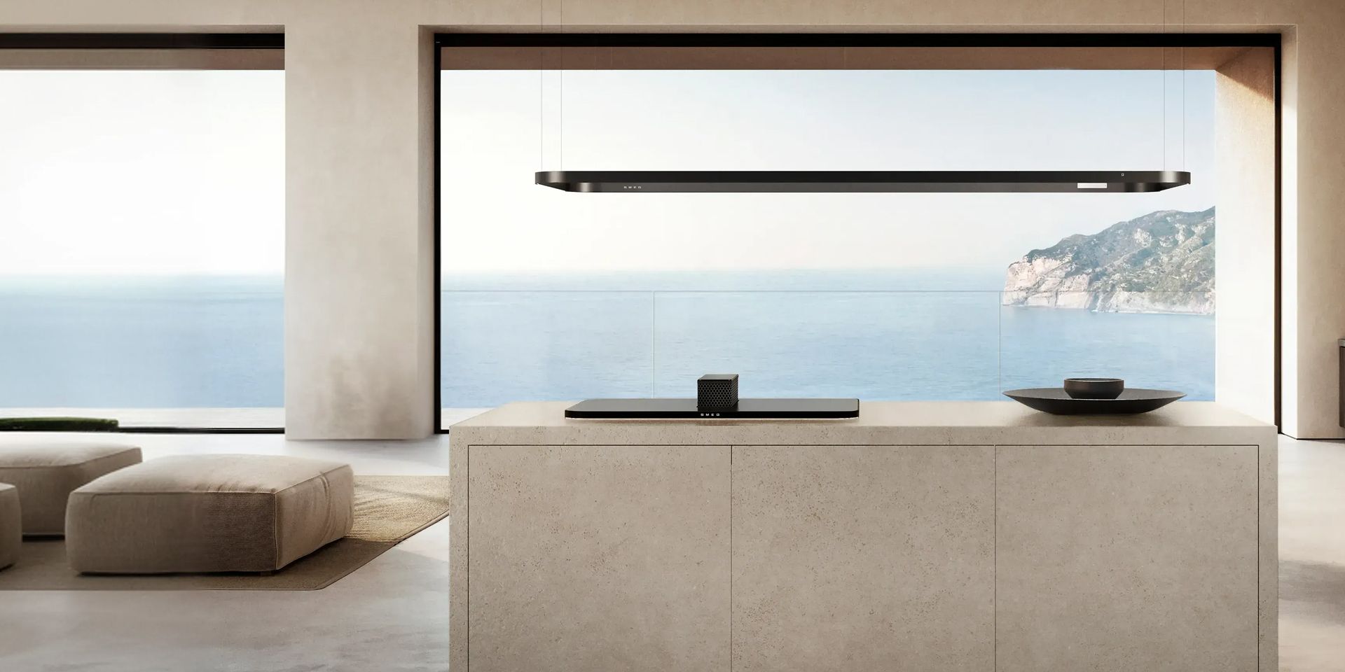 A sandy-beige stone kitchen island with a raised black hob and a black lighting rail, overlooking a blue ocean.