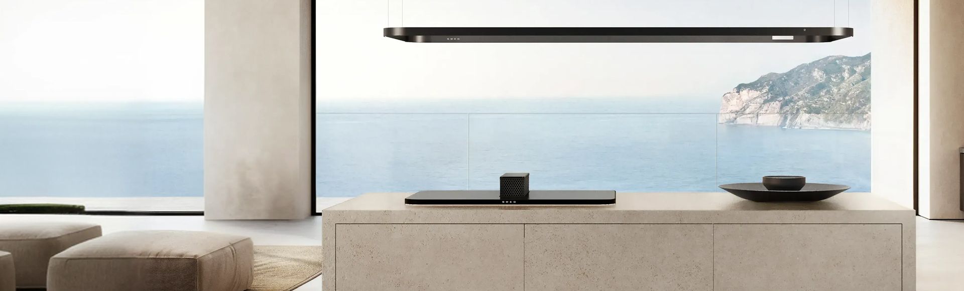 Un'isola da cucina in pietra color sabbia-beige con piano cottura nero rialzato e binario di illuminazione nero, con vista sull'oceano blu.