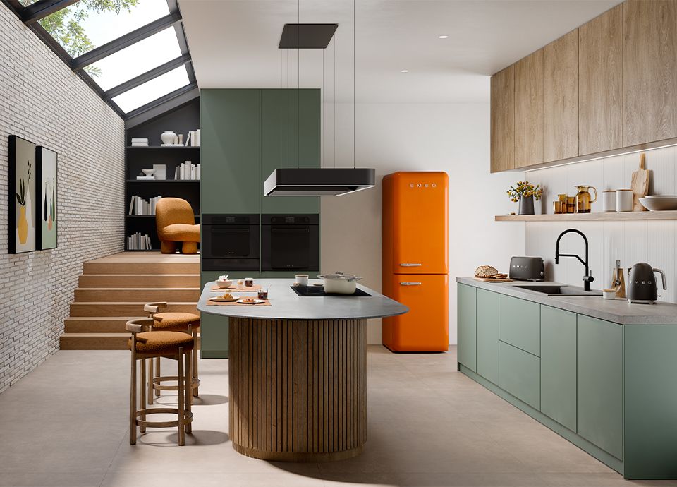 Modern kitchen featuring sage green cabinetry, a curved wooden slat kitchen island, and an orange Smeg FAB refrigerator. The space includes a skylight, white brick wall, and a raised seating area in the background.