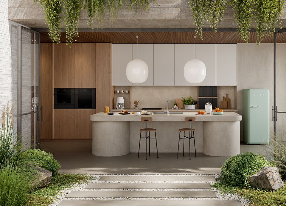 A modern kitchen featuring a curved beige stone island and wood-paneled wall with dual black ovens. A mint green retro refrigerator stands to the right, and the space opens directly to a lush green outdoor patio.