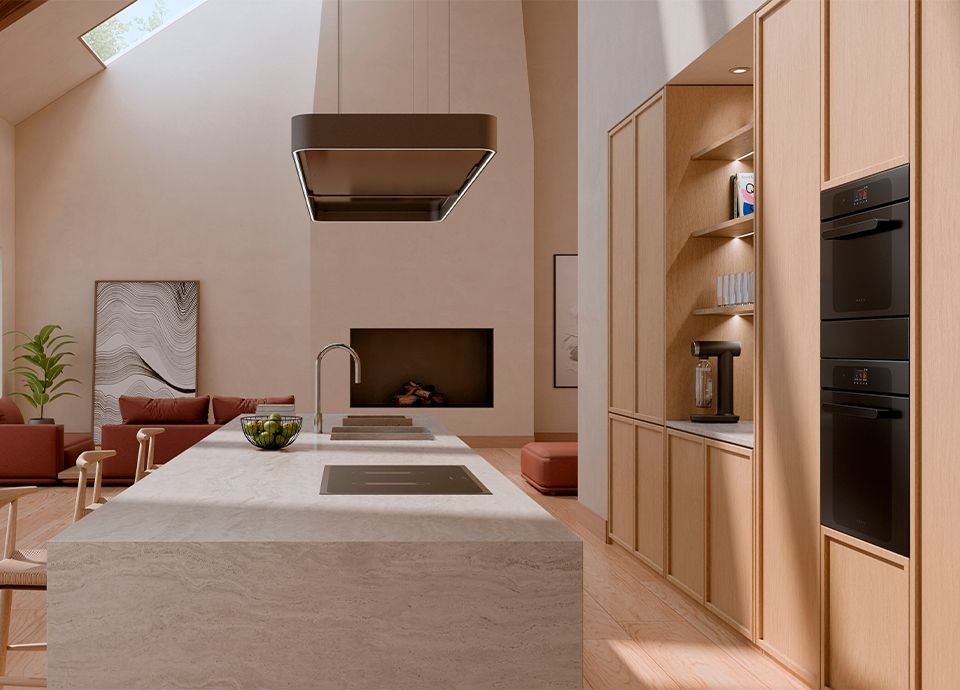 A minimalist kitchen and living area featuring a large light-grey marble island, light wood cabinetry with built-in black ovens, and a neutral-toned living space with a fireplace in the background.