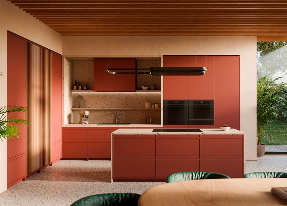 A modern minimalist kitchen featuring matte terracotta-red cabinetry, a large kitchen island with a cream countertop, and a wood-slat ceiling. The space includes built-in black ovens.