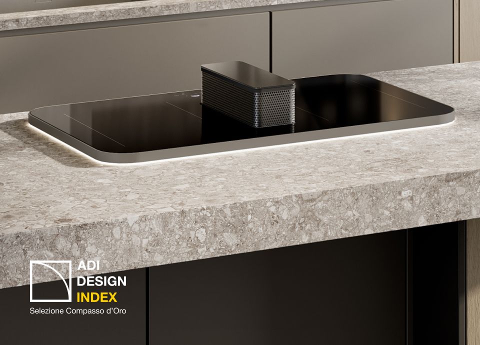 Black induction cooktop with a central integrated downdraft extractor, installed in a kitchen island with a light stone countertop; “ADI Design Index 2026” logo visible.