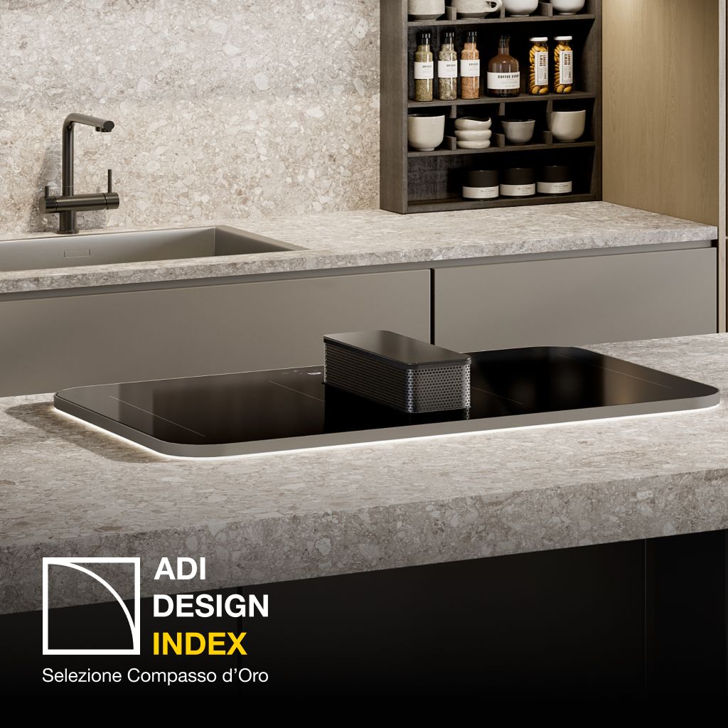 Black induction cooktop with a central integrated downdraft extractor, installed in a kitchen island with a light stone countertop; “ADI Design Index 2026” logo visible.