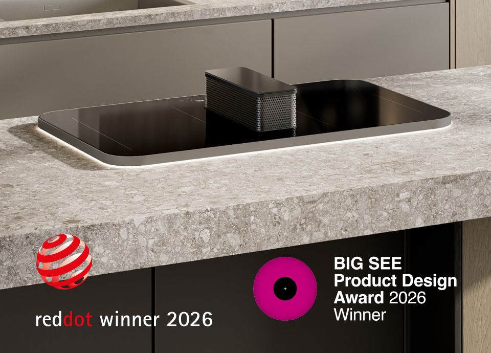 Black induction cooktop with a central integrated downdraft extractor, installed in a kitchen island with a light stone countertop; “Red Dot Winner 2026” logo visible.