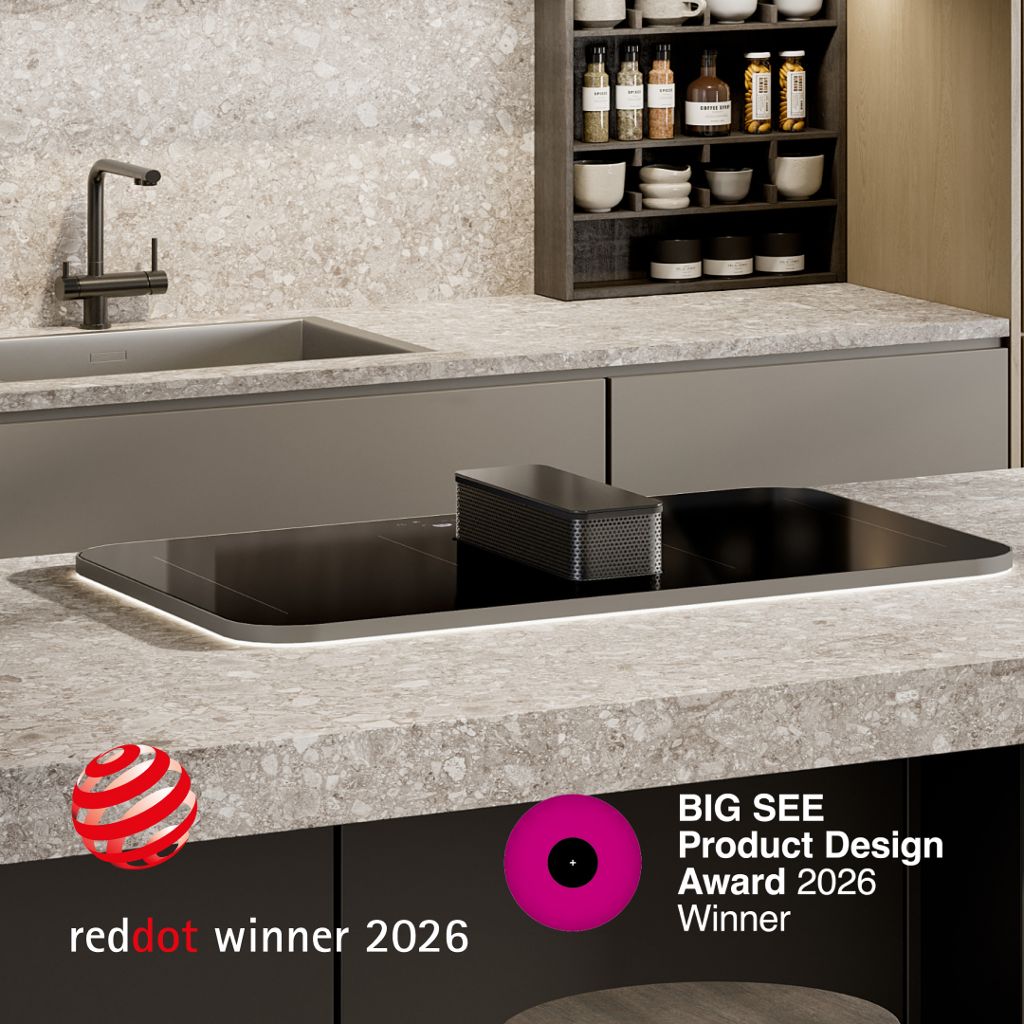 Black induction cooktop with a central integrated downdraft extractor, installed in a kitchen island with a light stone countertop; “Red Dot Winner 2026” logo visible.